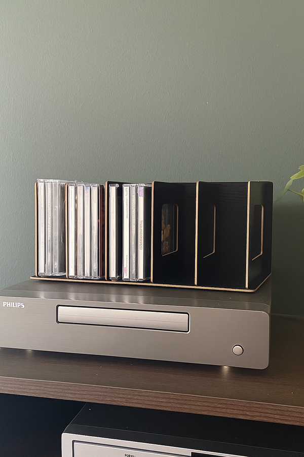 Een zwarte houten cd-houder, deels gevuld met cd's, staat bovenop een zilverkleurige Philips cd-speler op een donker meubel.