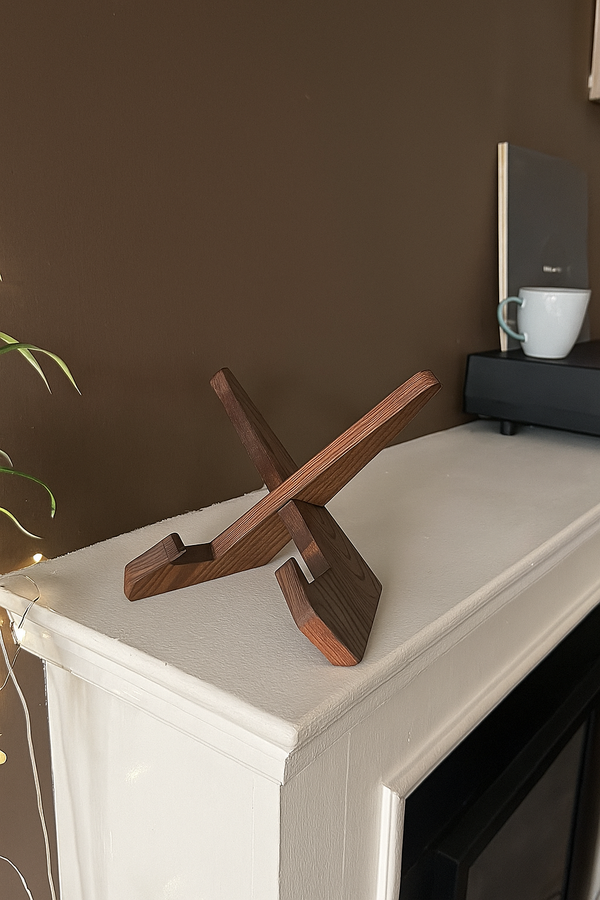 Een donker houten, x-vormige lp-houder staat leeg op een witte schouw in een gezellige woonkamer met een bruine muur.