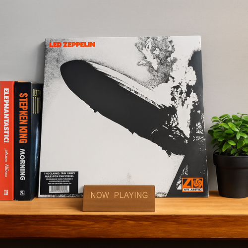 Een houten 'Now Playing' lp-standaard toont de iconische albumhoes van Led Zeppelin I, staand op een houten plank naast een stapel boeken en een groene plant.